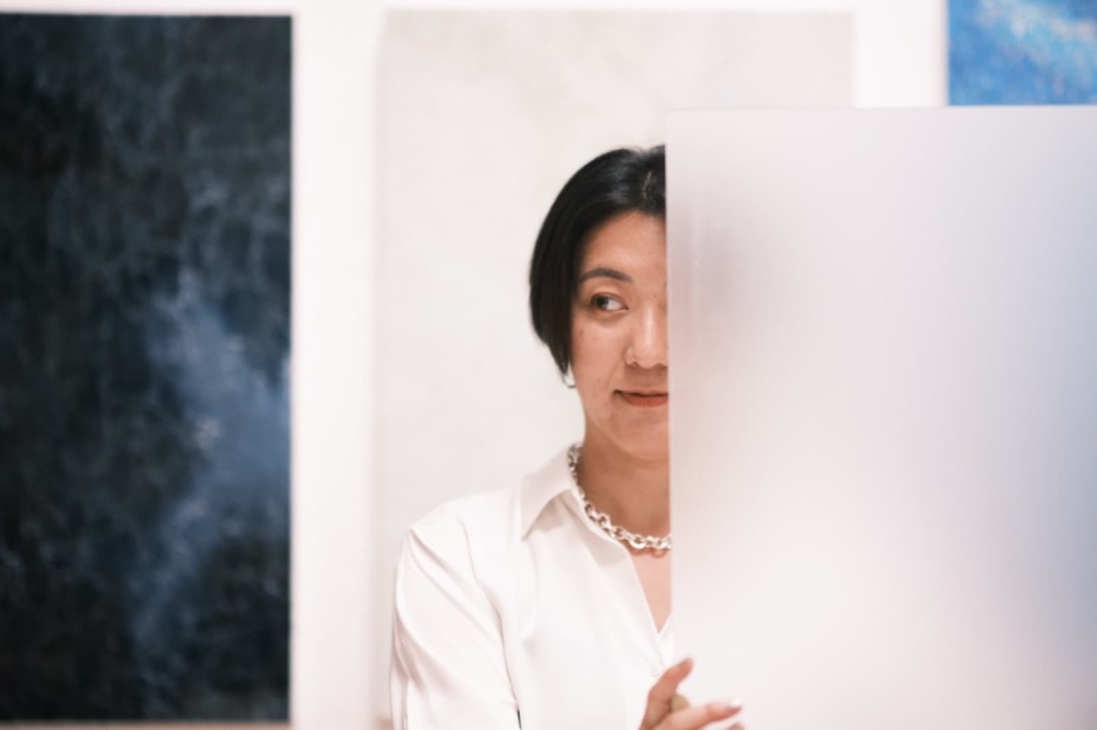 Half a face behind a translucent panel, one eye visible, paintings on the wall behind