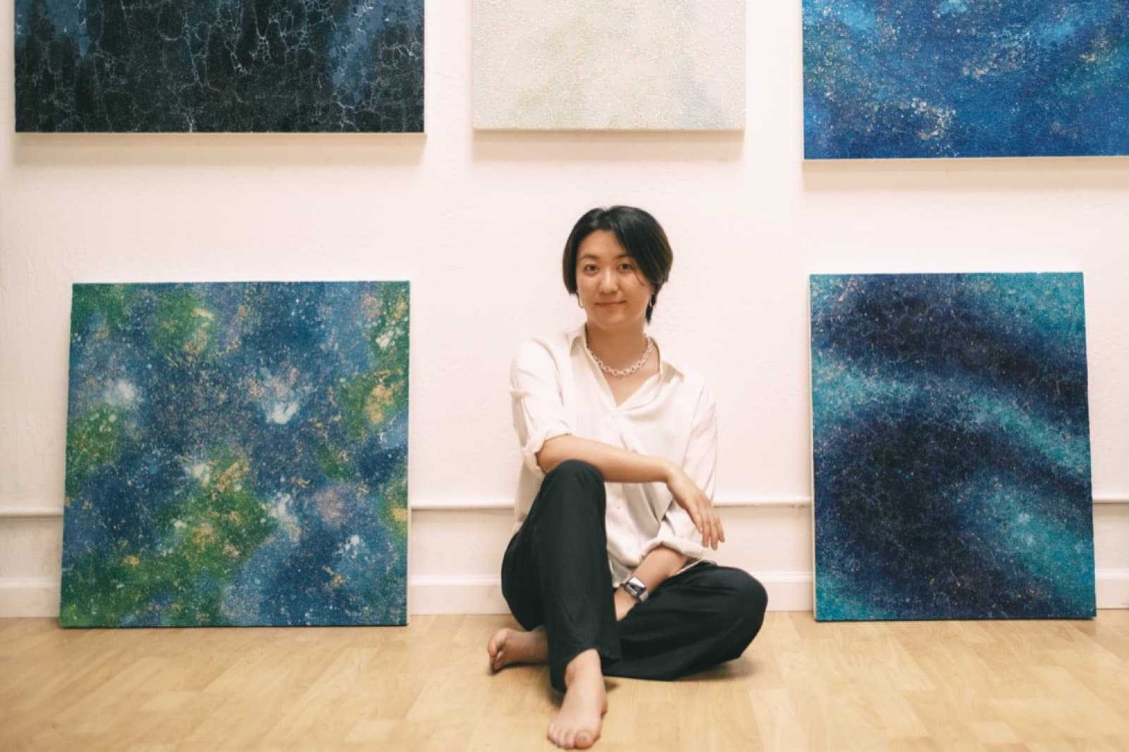 An artist sitting barefoot on a wood floor among her large blue and white paintings