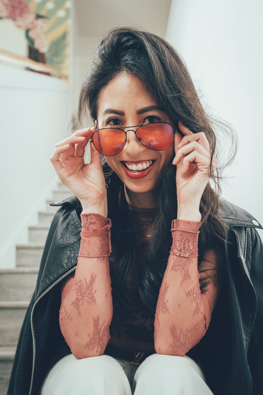 A woman on a staircase lifting tinted sunglasses, laughing toward the camera