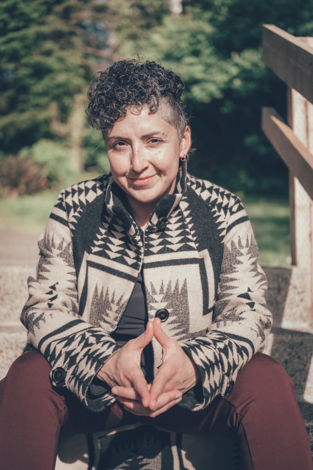 A woman seated outdoors in a patterned jacket, hands clasped, sunlight across her face