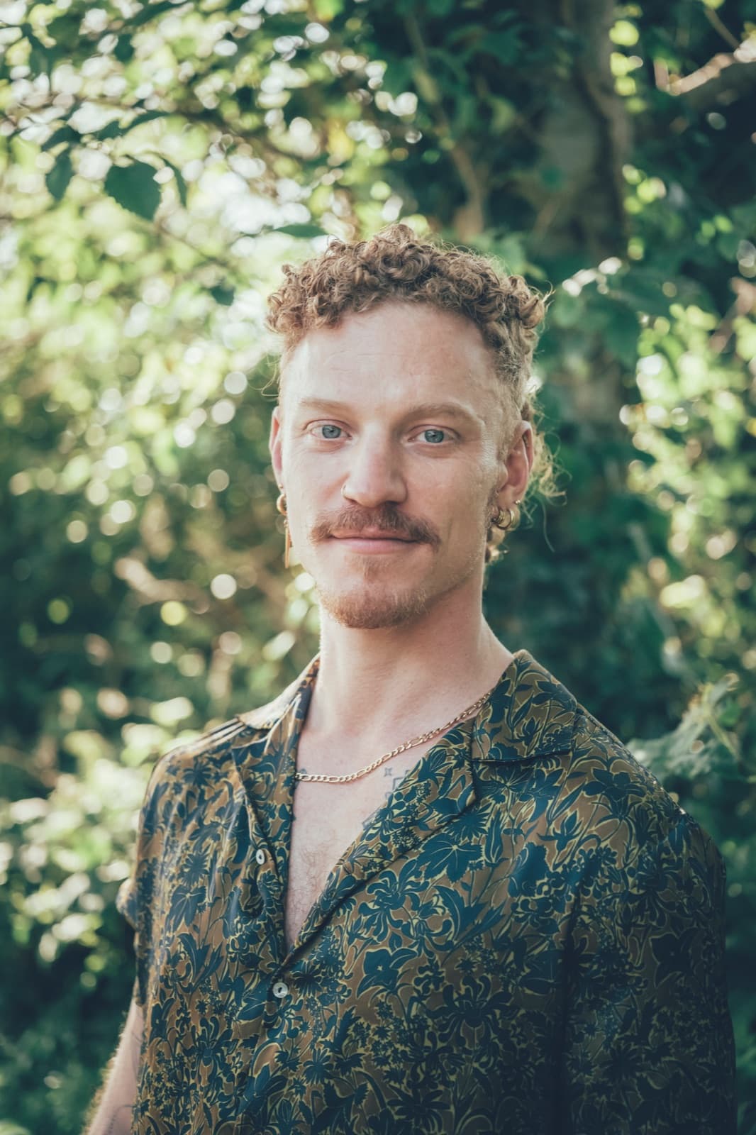 A man in a patterned shirt photographed close in dappled green light, looking straight to camera