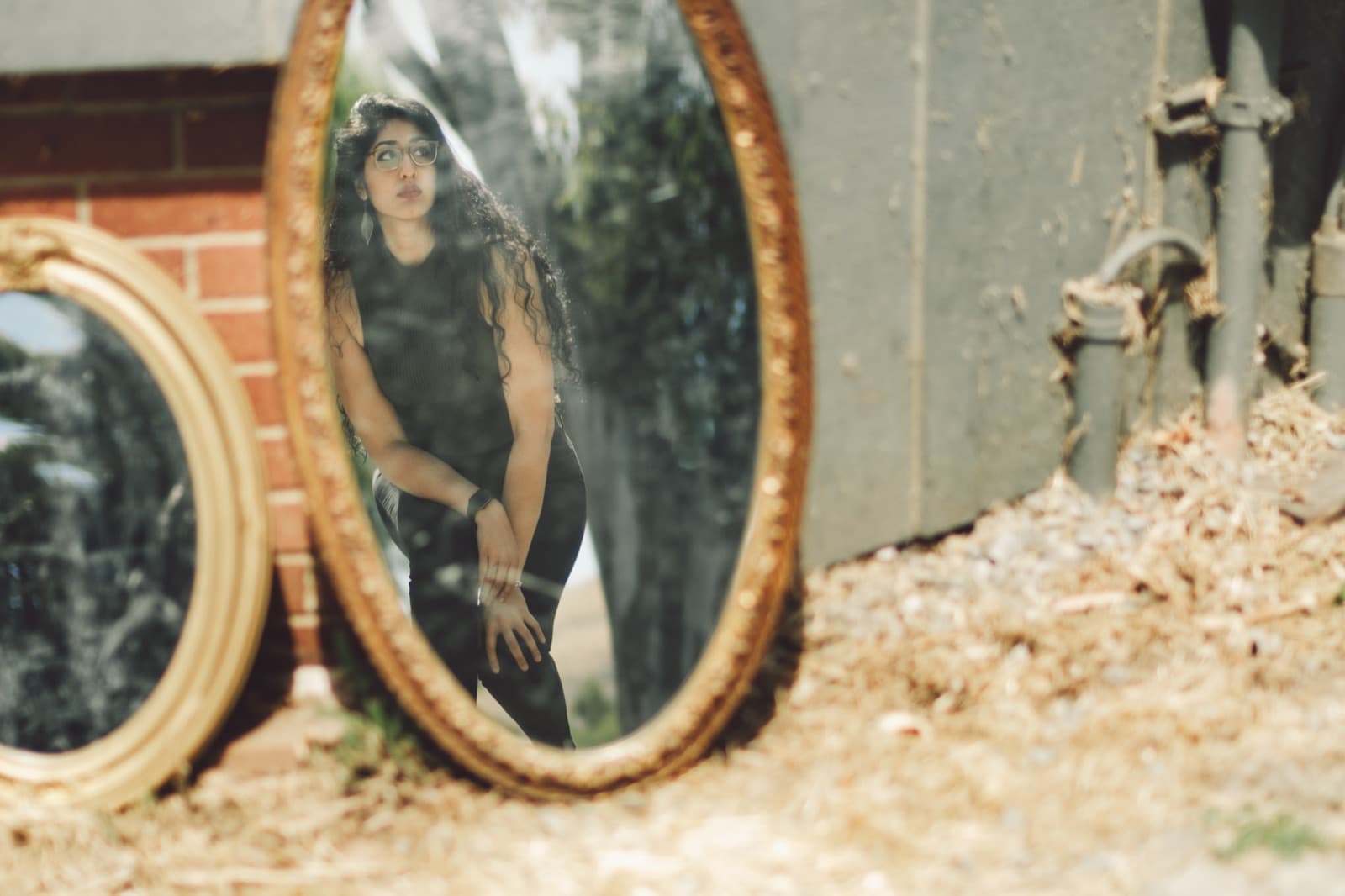 Two mirrors leaning against brick and corrugated metal, a woman visible only in the reflection